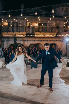 outdoor first dance at night on the stone terrace at Villa Antonia, bride and groom dancing hand-in-hand with the bride's flowing white gown billowing in motion and the groom in a dark navy three-piece suit with white boutonniere, warm Edison string light